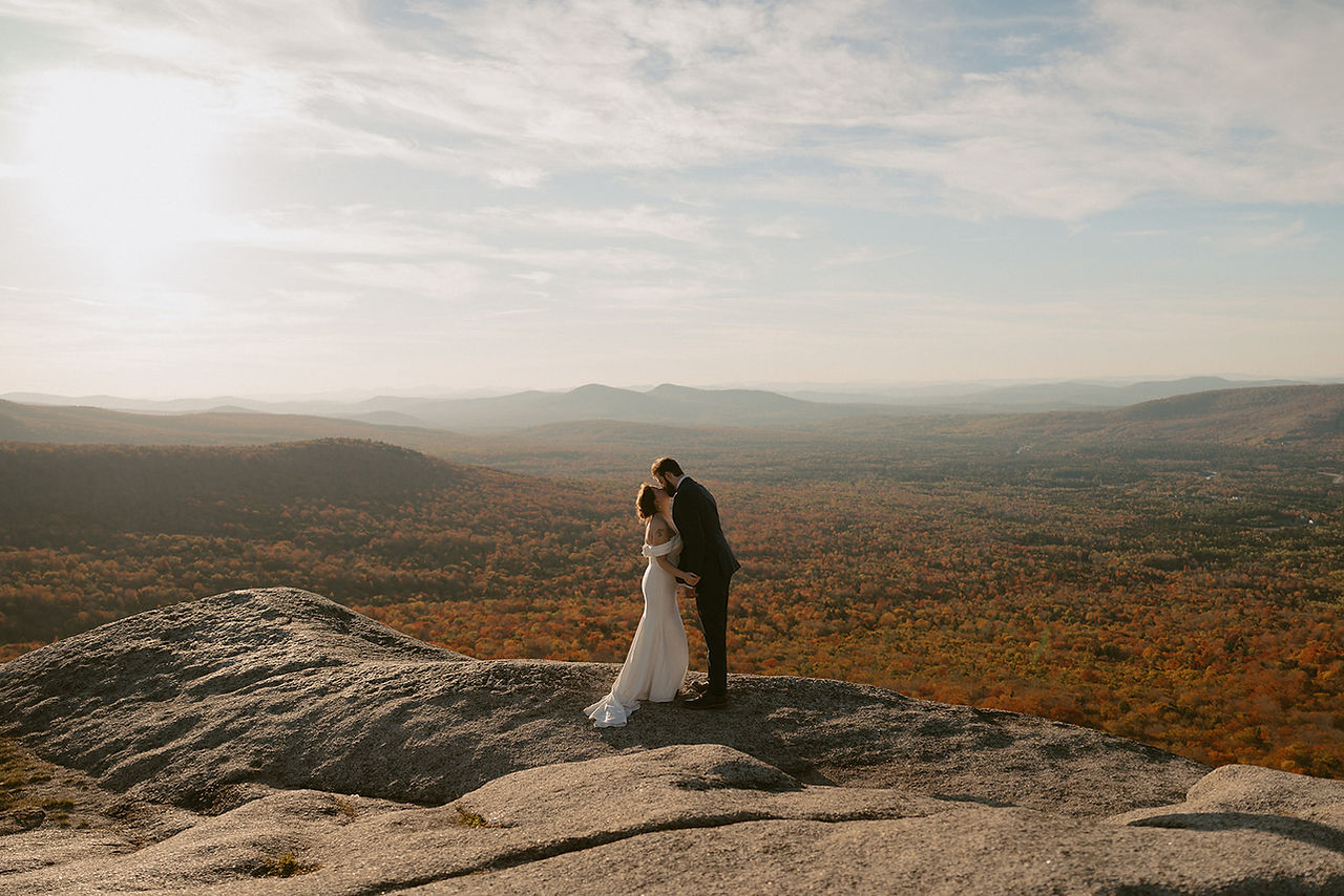Couple exploring and enjoying sunset from a mountain top for Crawford Notch elopement day in the White Mountains for an adventure elopement. The mountains behind them are in peak foliage in nh and the sun is golden as it sets.
