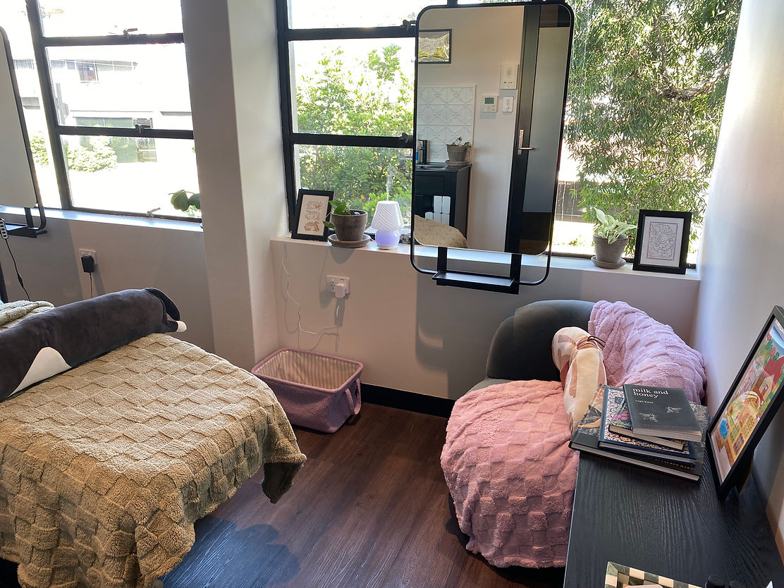 Bright, cozy room with large windows, a beauty bed with a plush green blanket and a long pillow, and a green chair with a purple plush blanket. Books, plants, and art adorn the room, creating a peaceful, inviting atmosphere.