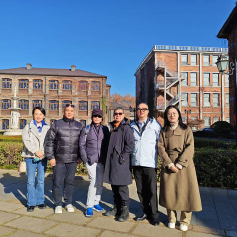 Group photo of six individuals standing outdoors on a paved walkway on a sunny day. From left to right: Tina, Washington, Ricky Ng, Mark, Panny, and a local Zhenjiang guide. Behind them are historic, multi-story brick buildings with distinctive arched windows and a central fountain.