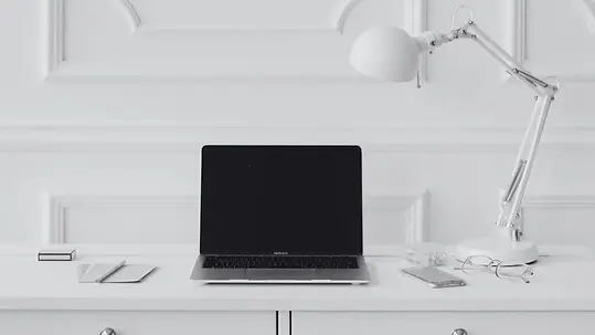 Minimal white desk setup with a laptop, desk lamp, notebook and glasses against a panelled