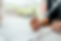 Hands sign a document with a silver pen on a table. Blurred green plants in the background. The mood is formal and focused.