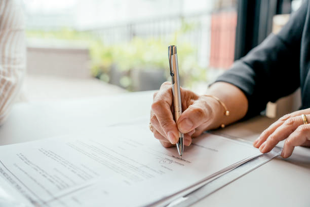 Hands sign a document with a silver pen on a table. Blurred green plants in the background. The mood is formal and focused.