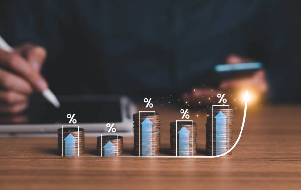 Stacked coins with percentage symbols and blue arrows indicate growth. A person writes and uses a phone in the blurred background. Mood is optimistic.