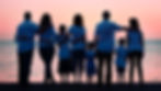 A group of people of various ages stand with their backs to the camera, overlooking a sunset over a body of water