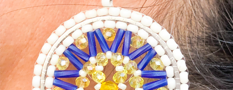 Pendientes Ruda de aro hechos a mano en estilo boho chic con cuentas Miyuki en tonos blanco, azul y amarillo, formando un diseño circular con detalles geométricos y cristales. Montados en ganchos de acero hipoalergénico, son ligeros pero impactantes, ideales para quienes buscan joyería artesanal vibrante con un toque bohemio y sofisticado.