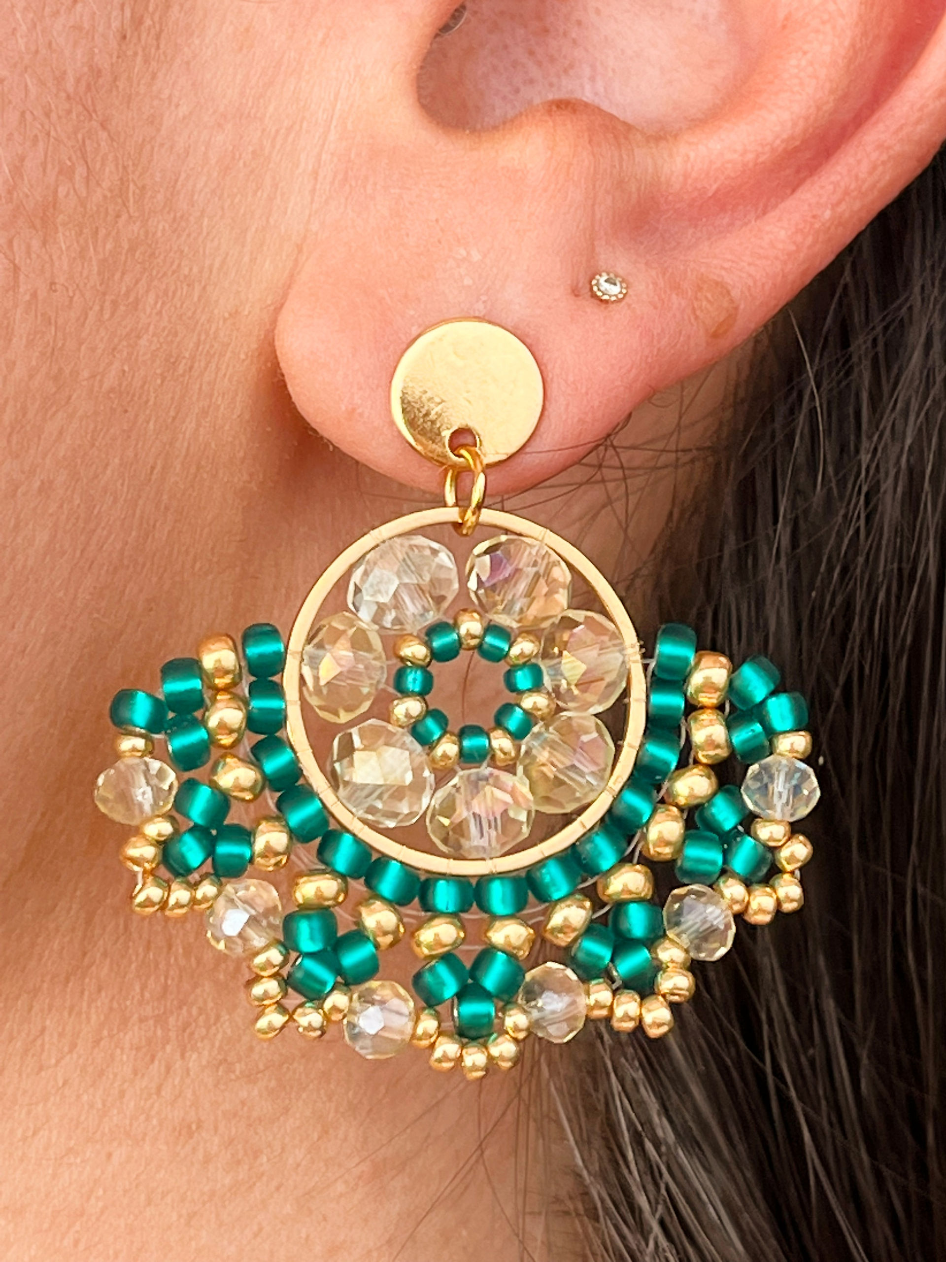 Pendientes Abba hechos a mano en estructura de acero dorado, con diseño circular y bordado de cuentas Miyuki en color verde botella, combinadas con bolitas de cristal checo transparente y dorado. Pieza llamativa y sofisticada, perfecta para complementar looks elegantes y originales con estilo artesanal.