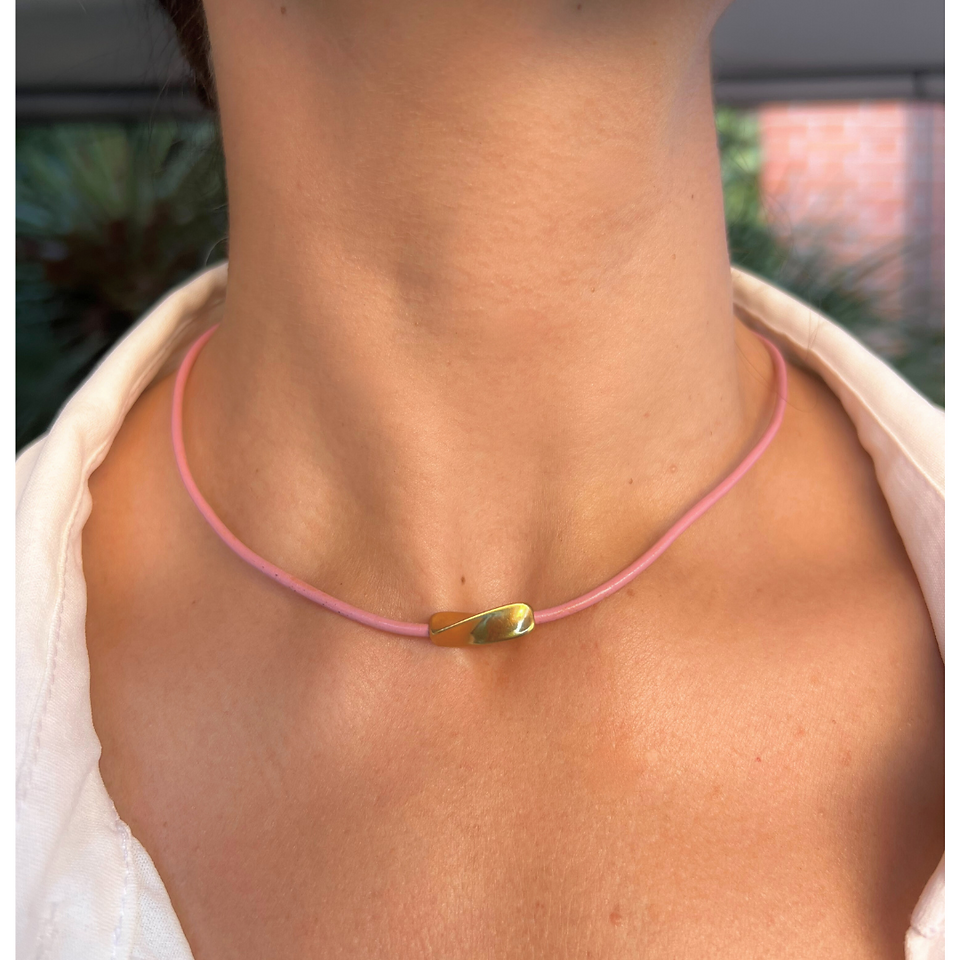 Collar Twist hecho a mano con un delicado cordón de cuero rosa y un elegante detalle central en acero dorado hipoalergénico. Su diseño minimalista y sofisticado lo convierte en un accesorio versátil, ideal para complementar cualquier look con un toque moderno y elegante.