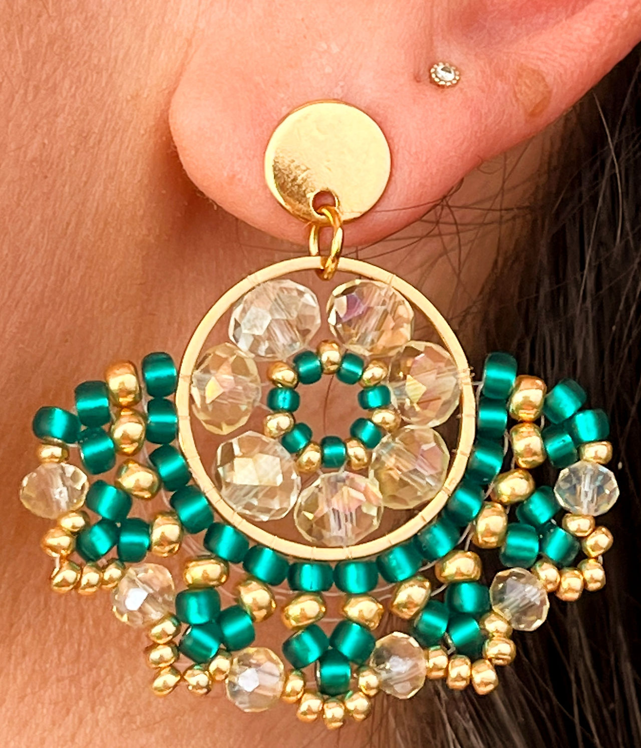 Pendientes Abba hechos a mano con cuentas Miyuki en tonos verde esmeralda y dorado, combinados con cristales checos transparentes que aportan brillo y sofisticación. Montados sobre una base de acero dorado hipoalergénico, su diseño en forma de abanico los convierte en un accesorio llamativo y elegante, ideal para ocasiones especiales y looks sofisticados.
