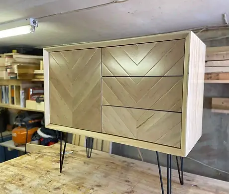 A handcrafted light ash wood cabinet in a woodworking shop, featuring a single large door on the left and four stacked drawers on the right, all showcasing a prominent natural wood grain.