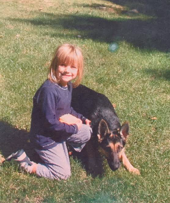 A young girl kneels next to a German shepherd in the grass.