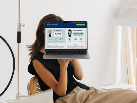 Woman sitting in a minimalist workspace holding a laptop in front of her face, displaying a blog post from the American Translators Association (ATA) explaining the difference between translators and interpreters