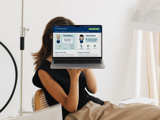 Woman sitting in a minimalist workspace holding a laptop in front of her face, displaying a blog post from the American Translators Association (ATA) explaining the difference between translators and interpreters