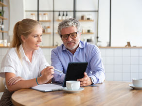 Young woman and older man review a tablet at a café table, with notebooks and coffee cups, in a focused, collaborative mood