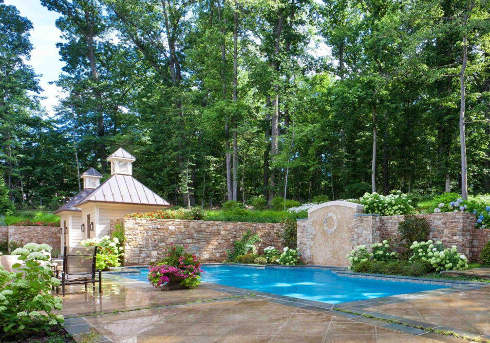 Backyard pool with stone walls, blooming flowers, and a small pool house beside tall green trees on a sunny day