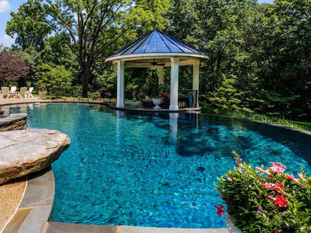 Blue backyard pool beside a white gazebo, surrounded by lush green trees and pink flowers on a sunny day