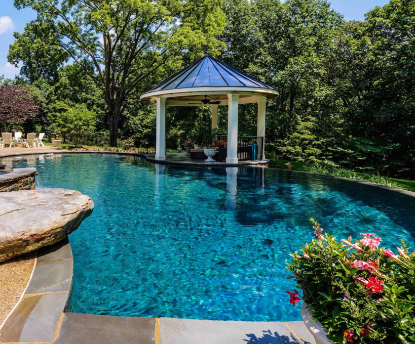 Blue backyard pool beside a white gazebo, surrounded by lush green trees and pink flowers on a sunny day
