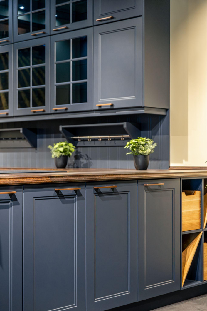 Modern blue-gray kitchen cabinets with wood countertop, potted plants, and open cubbies under warm lighting.