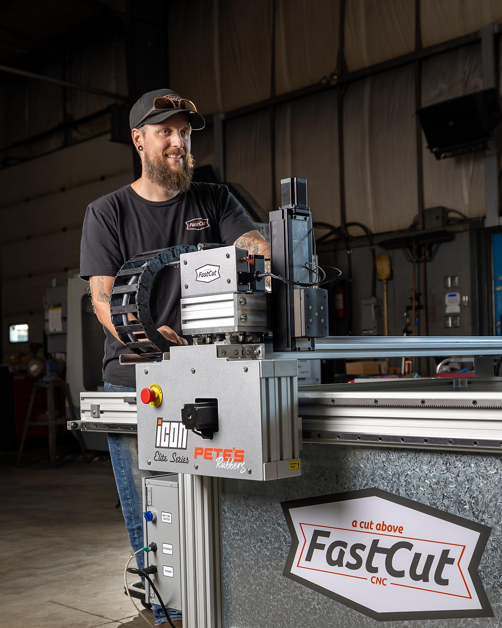 Smiling bearded man operates a FastCut CNC machine in a workshop, with large machine rails and logos visible.