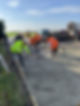 Workers in bright orange and yellow vests spread fresh concrete on a rural road