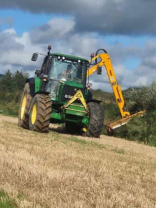 Green tractor with a yellow mechanical hedge cutting attachment working on a farm