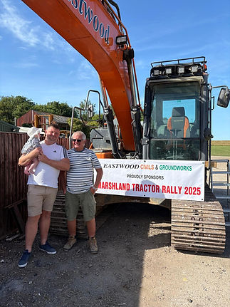 A man holding a baby and another man stand smiling in front of a large orange excavator with a banner reading, "Marshland Tractor Rally 2025."