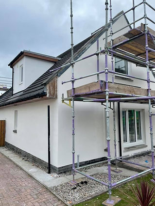 A small house with white plaster walls and a black-tiled roof is partially surrounded by scaffolding