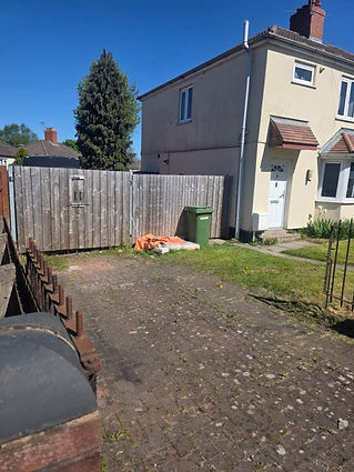 A beige two-story house with a fenced yard under a clear blue sky. The yard has a green bin and scattered bags