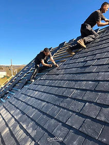 Roofer working on a steeply pitched roof installing dark grey roof slates under a clear sky
