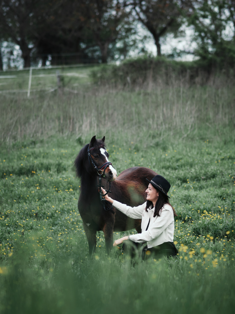 Frau mit Pferd sitzt im Gras, sie lächelt, Pferd und Frau sind sich zugewandt