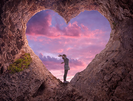 Woman standing in a heart-shaped cave, reaching towards a sunset sky.