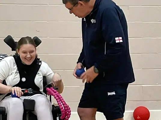 A young lady sat in her wheelchair laughing. She has blonde hair, which is tied back. She is wearing a short sleeved white t-shirt under her black wheelchair chest harness. The Boccia coach is stood on the right talking to the young lady. He is holding a blue Boccia ball and a dark navy half-zip jumper with white detail and the England flag on the left upper arm.