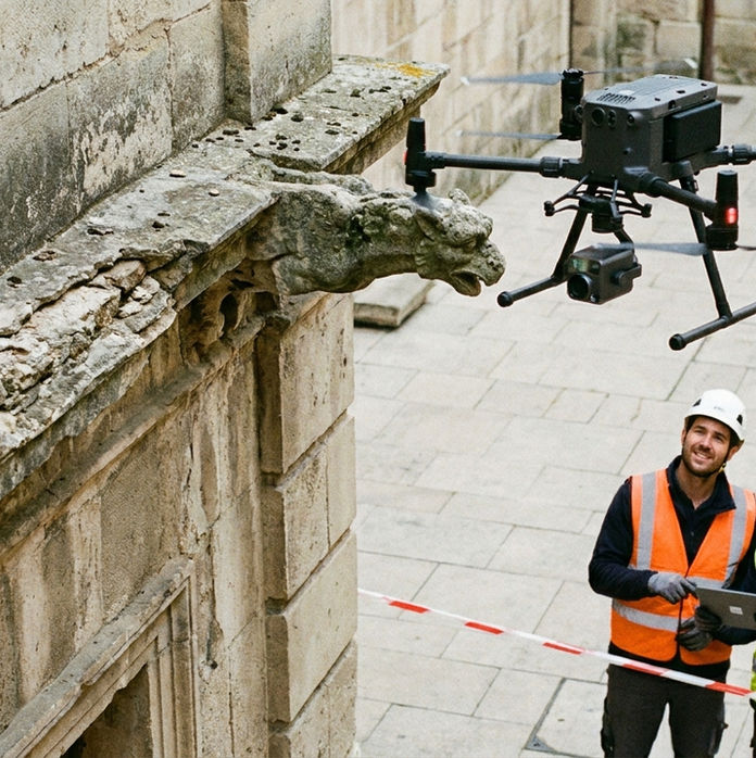 Deux ouvriers en gilets orange et vert contrôlent un drone près d'une gargouille en pierre, dans une cour pavée. Atmosphère professionnelle.