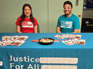 Nick Moncada (right) and a fellow JFAI team member sit behind an outreach table covered with immigration resource materials during a community event hosted by Justice for all Immigrants.