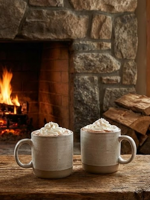 Zwei Tassen heißer Kakao vor einem Kaminfeuer.