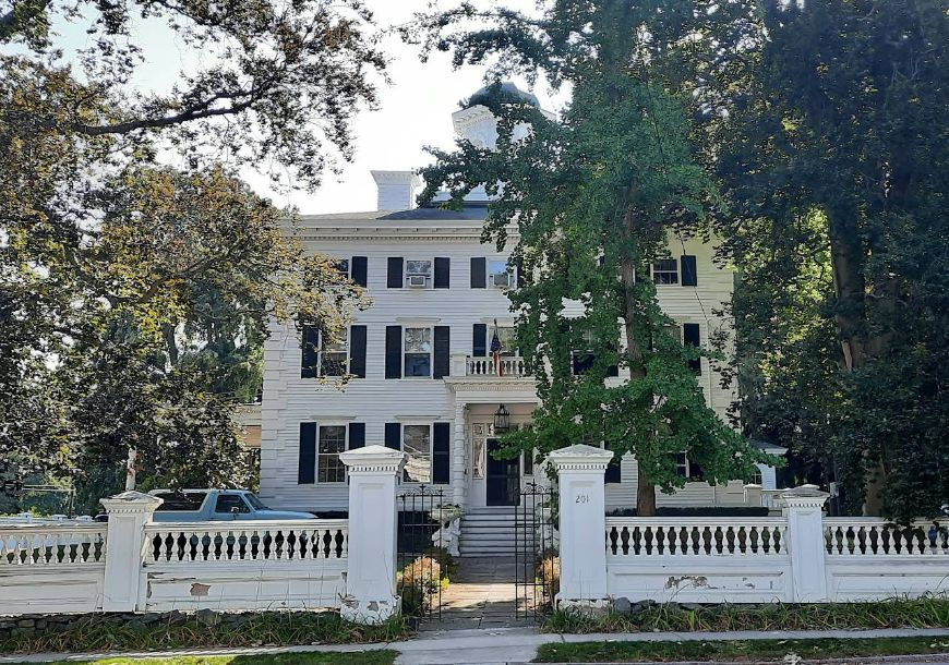 White three-story house with black shutters and a wraparound porch, surrounded by tall trees and a white fence. House number 201 visible.
