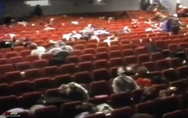 People are lying motionless on red theater seats in a dimly lit auditorium, creating a tense and unsettling atmosphere.
