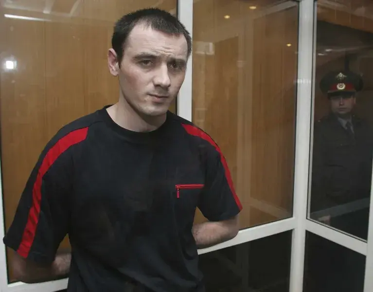 A man in a black and red shirt stands with hands behind his back in a glass enclosure, serious expression. A guard in uniform is visible outside.