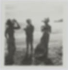 Three women in swimsuits and hats stand on a sunny beach. One adjusts her hat. They wear patterned skirts, with ocean waves in the background.