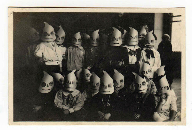 Children wearing ghost masks pose in a dimly lit room. The black-and-white image has an eerie, vintage feel.