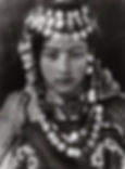 A person wears traditional attire adorned with coins, a headdress, and floral patterns. The mood is serious and the setting is neutral.