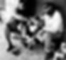 Two men sit on a sidewalk with boxes, sharing a meal. One holds a spoon. They're engaged in conversation. Visible text: "BAFAS." Mood: calm.