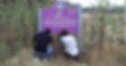 Two people kneeling, reading a bullet-riddled, purple sign labeled "River" in a grassy area. They appear contemplative.