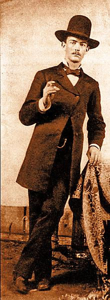 Man in vintage suit and hat poses with a book, leaning on a patterned chair. Background is plain, sepia tone adds historic feel.