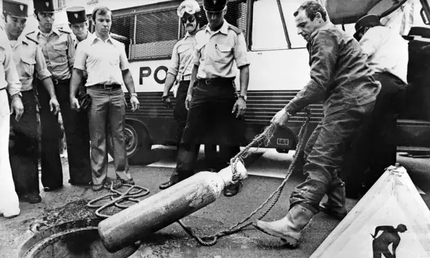 Police officers and a man in uniform lift a cylinder from a manhole. A police van is in the background. The scene appears tense and focused.