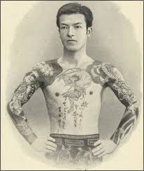 Man with full tattoo sleeves and chest tattoos stands confidently with hands on hips. Black and white photo, neutral background.