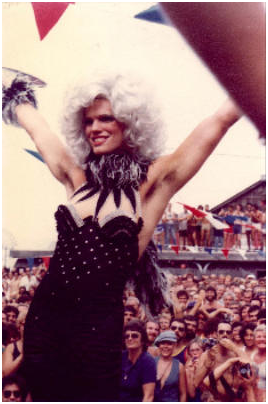 Drag queen with white hair, black sequin outfit celebrating at outdoor festival.