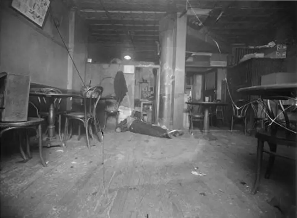 Interior of an old cafe with empty tables and chairs, a person lying on the floor. Dim lighting creates a somber mood. Black and white.