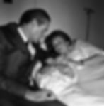 Svetlana Stalin in a hospital bed holds a newborn Chrese Evans, gazing lovingly at a man in a suit beside her. The room is dimly lit, creating a tender mood.