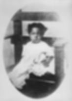 Child sitting in an ornate chair, wearing a white dress with lace. The photo has a vintage feel with a soft, serious expression.