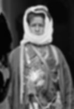 A person in traditional attire, wearing a headscarf and bullet bandoliers across the chest. Dark background, serious expression. Black and white image.